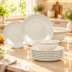 A speckled off-white ceramic dinnerware set arranged on a marble kitchen countertop, including plates, bowls, and a mixing bowl, with a bright kitchen background.