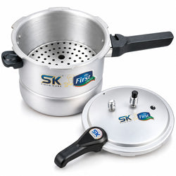 A stainless steel pressure cooker set on a white background, featuring a main cooker with a perforated steaming insert, a separate lid, and black handles. The cooker and lid display the SK 'The First' brand logo.