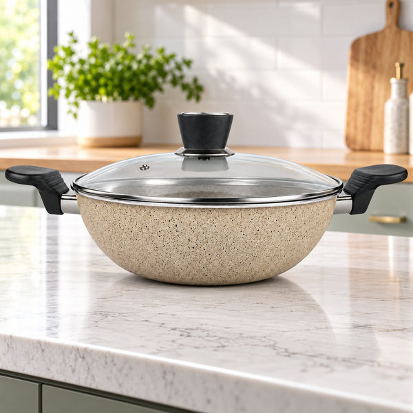 A speckled beige nonstick pan with a clear glass lid and black handles sits on a white marble kitchen countertop.