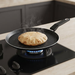 A black non-stick frying pan with a handle sits on a lit blue gas stove burner in a kitchen setting, with a round flatbread cooking inside.
