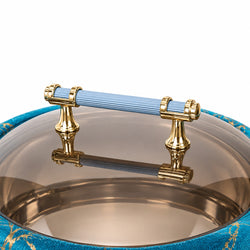 A close-up of a round glass lidded container with a transparent smoky glass top, a ribbed light blue handle, and ornate gold metal hardware, set against a white background.
