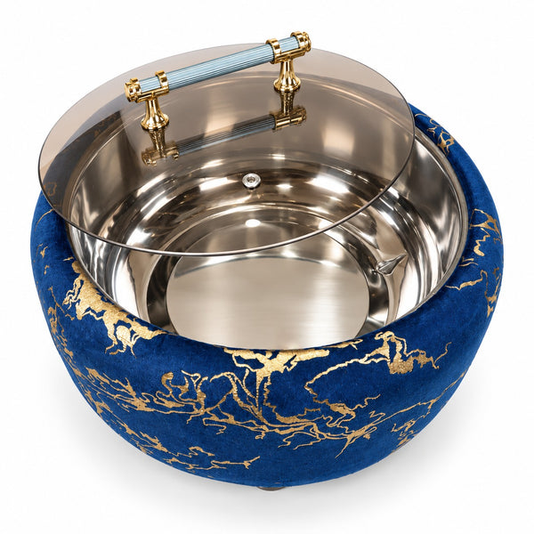 A round blue velvet stool or ottoman with gold dragon-style patterning, featuring a polished stainless steel bowl-like insert and a clear glass lid with a light blue handle and gold hardware, shown on a white background.