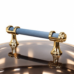 A close-up of a kitchen pot or pan handle with a ribbed blue grip and polished gold-tone metal ends, resting on a reflective metallic surface against a white background.