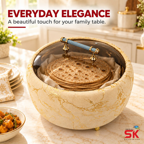 An elegant round tabletop steamer or warmer with a cream marble-patterned exterior and gold detailing, shown on a marble countertop in a bright kitchen. Inside, flatbread or tortillas are stacked on a liner under a clear lid with a blue handle.